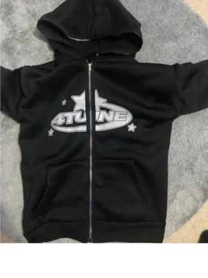 Svart hoodie med 4TUNE-tryck - Snygg svart hoodie med dragkedja och stor huva. På bröstet finns ett stort silverfärgat 4TUNE-tryck med stjärndetaljer. Hoodien har två rymliga fickor framtill och är gjord i mjukt material som känns skönt mot huden. Perfekt för en avslappnad streetwear-look.
