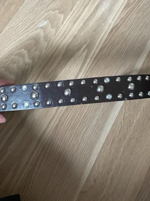 Brunt skärp med nitar i skinn - Snyggt svart/brunt skärp i skinn med silverfärgade nitar i olika storlekar längs hela bandet. Skärpet har ett klassiskt spänne i silver och passar perfekt för att ge outfiten en cool touch. Lädret har åkt av lite vid de små hålen