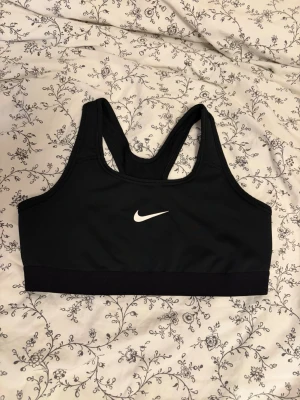(RESERVERAD!!!)Svart sport-bh från Nike - Svart sport-bh från Nike med klassisk racerback-design och vitt Nike-logga framtill. Tillverkad i stretchigt och fukttransporterande material som passar perfekt för träning och sportaktiviteter. PASSAR XS-S