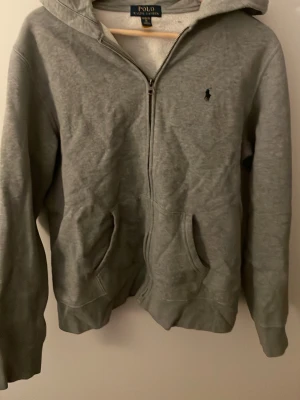 Grå hoodie från Polo Ralph Lauren - Snygg grå hoodie från Polo Ralph Lauren med dragkedja framtill och två fickor. Klassisk broderad logga på bröstet och mjukt, fleecefodrat innerfoder. Perfekt för en avslappnad och stilren look. Huva med dragsko för extra komfort.