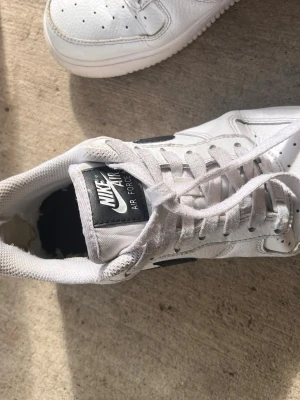 Nike Air Force 1 vita sneakers - Klassiska Nike Air Force 1 sneakers i vitt läder med svarta detaljer och Nike-logga på sidan och hälen. Skorna har perforerad tå för ventilation, platt vit sula och lågt skaft. Perfekta för en stilren och sportig look.