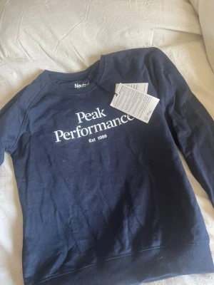 Mörkblå Peak Performance sweatshirt - Mörkblå sweatshirt från Peak Performance med vit logga och text på bröstet. Klassisk rund halsringning och ribbade muddar vid ärmslut och nederkant. Perfekt för en avslappnad och sportig stil.