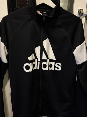 Svart Adidas träningsjacka  - Snygg svart träningsjacka från Adidas med vit logga och vita detaljer på ärmarna. Jackan har hel dragkedja framtill och hög krage. Perfekt för träning eller chill. Tillverkad i ett mjukt och funktionellt material.Youth Xl . Skriv för frågor eller buda