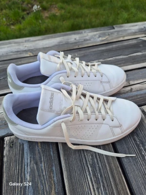 Vita Adidas sneakers med grå detalj - Fräscha vita sneakers från Adidas med klassisk låg profil och diskreta grå detaljer på hälen. Skorna har perforerade sidor, vita skosnören och mjuk vadderad insida. Perfekta för en clean och stilren look.