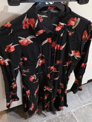 Svart blommig klänning från Zara - Svart klänning från Zara med röda och vita blommor, långärmad och med knappar framtill. Klänningen har klassisk krage och är tillverkad i ett lätt tyg som ger en snygg siluett. Perfekt för dig som gillar blommiga mönster och stilren design.