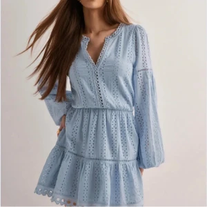 Ljusblå broderad kortklänning - Söt ljusblå kortklänning med broderade detaljer och v-ringning. Klänningen har långa ärmar med lätt puff och resår i midjan för en feminin siluett. Tillverkad i bomull, perfekt för varma dagar och somriga tillfällen.