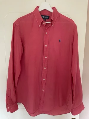 Korall röd linneskjorta från Ralph Lauren - Snygg korall röd linneskjorta från Ralph Lauren med klassisk button-down krage och broderad logga på bröstet. Skjortan har lång ärm och custom fit-passform, perfekt för en avslappnad men stilren look.