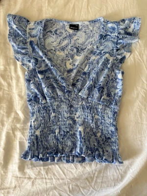Blåvit omlottopp från Gina Tricot - Supersöt blåvit omlottopp från Gina Tricot med volangdetaljer på axlar och nederkant. Toppen har v-ringning, korta ärmar och smockad midja för en snygg passform. Perfekt till jeans eller kjol för en fräsch look.