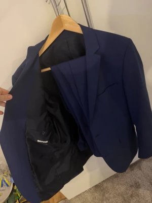 Mörkblå kostym med kavaj och byxor - Snygg mörkblå kostym med klassisk kavaj och matchande byxor. Kavajen har två knappar, smala slag och innerficka. Perfekt för dig som vill ha en stilren och modern look. Passar till skjorta och sneakers eller finskor.
