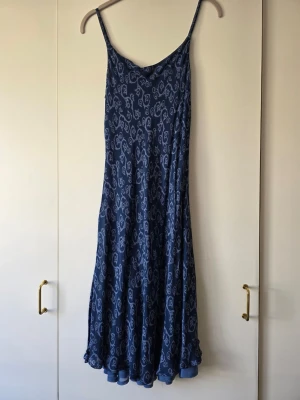 Blå sommar klänning med liknande paisleymönster - Snygg blå långklänning med tunna axelband och paisleyinspirerat mönster i ljusare blå nyans. Klänningen har en flowy passform och är perfekt för varma dagar. Materialet känns mjukt och lätt, tviskos.