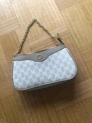 Gucci beige axelremsväska med kedja - Snygg beige handväska från Gucci med klassiskt GG-mönster, guldfärgad kedjerem och detaljer i ljusbeige läder. Väskan har dragkedja upptill och ikonisk GG-logga framtill. Perfekt för dig som gillar lyxiga accessoarer och vill sticka ut.