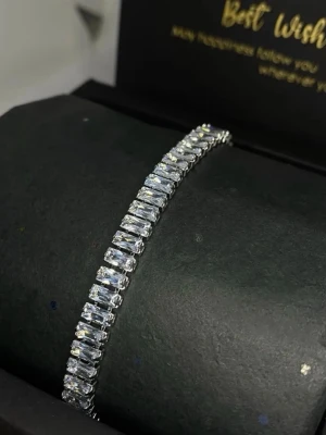 Silverfärgat tennisarmband med baguette stenar - Ett stilrent tennisarmband i silverfärg med rektangulära, gnistrande baguette formade stenar som sitter tätt i rad. passar till många olika stilar. Väldigt VVS, skiner fint! Funkar för kvinnor & män
