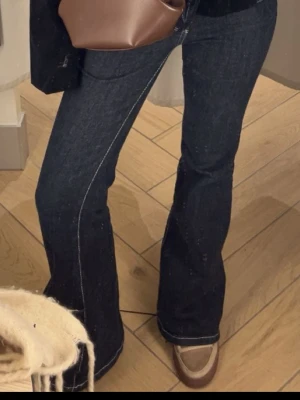 Mörkblå bootcut jeans med kontrastsömmar - Snygga mörkblå bootcut jeans med vita kontrastsömmar och klassisk femficksdesign. Jeansen har låg midja och utsvängda ben, vilket ger en retro och trendig vibe. Perfekta att styla med sneakers eller boots.