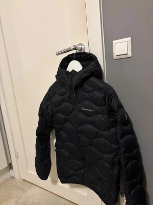 Svart dunjacka från Peak Performance Small Herr - Storlek S Herr ! Snygg svart dunjacka från Peak Performance med vågformat mönster och huva. Jackan har dragkedja framtill och logga på bröstet. Perfekt för kalla vinterdagar och riktigt skön att bära. Klassisk modell som funkar till det mesta. Har slitits upp lite på baksidan längst ner vid ryggen men inget som synd mycket. Annars är den i ett väldigt bra skick men det var det lilla som fick den att hamna i acceptabelt skick!