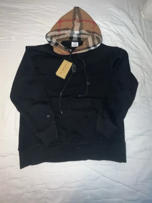 Burberry hoodie - Snygg svart hoodie från Burberry med den klassiska rutiga huvan i beige, rött och svart. Jätte bra skick storlek M passar även S. Pris kan diskuteras!