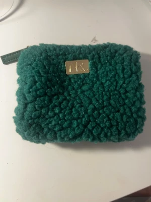 Grön fluffig plånbok med gulddetalj - En mysig, fluffig grön plånbok med en rektangulär form och guldfärgad metallbricka framtill. Plånboken har dragkedja och är klädd i ett mjukt, teddy-liknande material som ger en unik och trendig look.