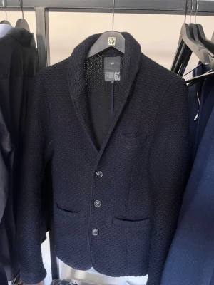 Stickad mörkblå kavaj  - Snygg mörkblå stickad kavaj från 66°North med tre knappar framtill, klassisk krage och fickor. Kavajen har en grovstickad struktur som ger en modern och avslappnad look. 