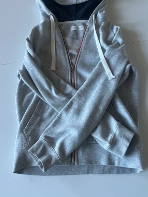 Grå zip-hoodie från Pier One - Snygg grå hoodie från Pier One med dragkedja och blå, röda detaljer runt dragkejdan . Mjuk insida och fickor framtill. Perfekt för en chill och avslappnad stil. Klassisk passform och enkel att matcha med det mesta.