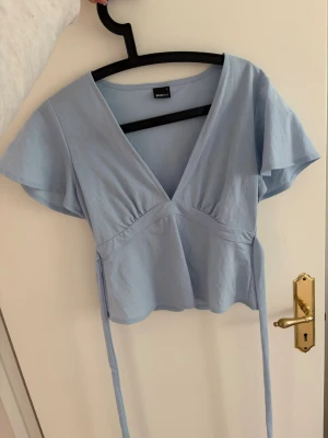 Ljusblå v-ringad topp Gina Tricot - Supersöt ljusblå topp från Gina Tricot med v-ringning och knytband i midjan. Toppen har korta, fladdriga ärmar. Kommer inte till användning och säljs därför! Den har inga defekter då den knappt är använd🫰skickar samma dag