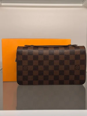 Brun handväska - Snygg handväska i klassiskt brunt mönster. Väskan har guldfärgade detaljer, dragkedja och är tillverkad i tåligt canvasmaterial med läderdetaljer. Perfekt accessoar för dig som gillar lyxig och tidlös design.
