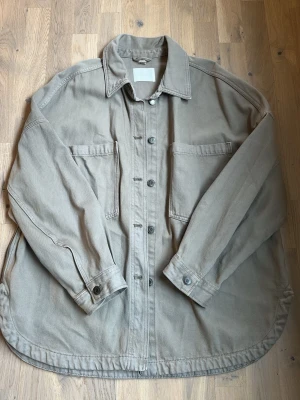 Beige jeansjacka med fickor - Stilren beige jeansjacka i kraftigt bomullstyg med två stora bröstfickor och knappar framtill. Skjortan har klassisk krage, långa ärmar med knapp vid manschett och avslappnad passform. Perfekt lager på lager-plagg för en casual look. Beige/grå färg.