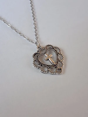 Silverfärgat halsband med hjärta och kors - Snyggt halsband i silverfärg med ett detaljerat hjärthänge och ett kors i mitten. Hänget har en pärlad kant och kedjan är tunn och stilren. Perfekt accessoar för dig som gillar unika smycken med symbolik.