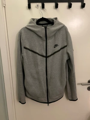 Grå Nike tech dress  - Snygg grå hoodie från Nike med hel dragkedja framtill och svart Nike-logga på bröstet. Byxor tillkommer. Hoodien har en huva och svarta detaljer längs bröstet och ärmsluten. Tillverkad i mjukt material som känns skönt mot huden. Perfekt för en avslappnad och sportig stil. Koftan och byxorna är i M 