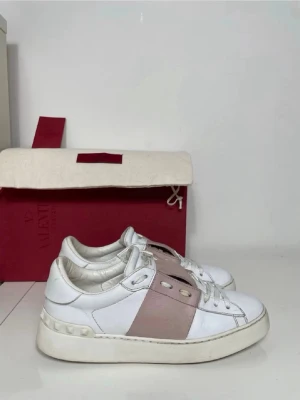 Valentino Garavani vita sneakers - Snygga vita sneakers från Valentino Garavani med bred rosa rem över mitten. Skorna har klassisk låg modell, vita snören och en chunky sula. Perfekta för dig som gillar exklusiv och trendig streetstyle.