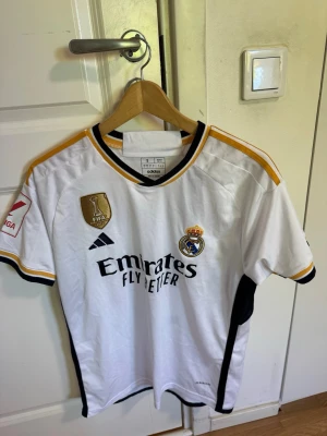 Real Madrid matchtröja Adidas S - Säljer en vit Real Madrid matchtröja från Adidas i storlek S. Tröjan har svarta och gula detaljer, klubbmärke, FIFA-märke och sponsortryck framtill. Slim fit-modell med kort ärm och broderade loggor. Perfekt för fotbollsfans!