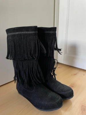Svarta mockastövlar med fransar - Snygga svarta stövlar i storlek 38 i mocka med dubbla rader fransar längs skaftet. De har en rund tå och grov sula som ger ett coolt uttryck. Perfekta för dig som vill sticka ut med din stil och gillar boho-vibbar.
