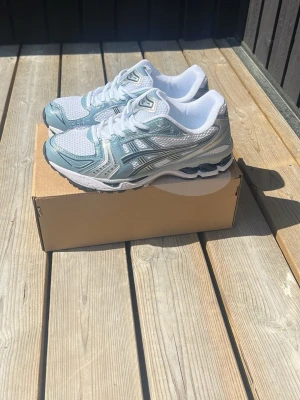 ASICS vita och blå sneakers - Snygga sneakers från ASICS i vitt och grått med klassisk mesh- och syntetdesign. Skorna har tydliga ASICS-loggor på sidorna, vadderad plös och häl samt robust sula för bra komfort. Perfekta för en sportig och trendig look.