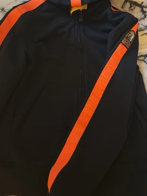 Svart hoodie med orange ränder - Cool svart hoodie från Parajumpers med dragkedja och huva. Har tydliga orange ränder längs ärmarna och axlarna samt ett broderat Parajumpers-märke på ena ärmen. Perfekt för en sportig och avslappnad stil.