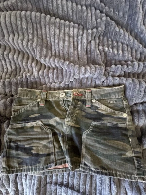 Militär kortkjol  - Snygg kortkjol från Urban outfitter i klassiskt militär-mönster. Kjolen har stora fickor fram och bak, bälteshällor och detaljer med kontrastsömmar. Perfekt för en cool och avslappnad stil.