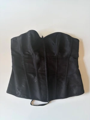 Svart satin korsett med snörning - Stilren svart korsett i satin med lätt glans och diskret mönster. Formpressad byst och markerade sömmar för snygg siluett. Snörning i ryggen och dragkedja framtill för enkel påtagning. 
