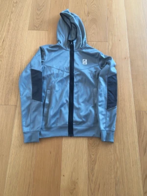 Ljusgrå hoodie med dragkedja från Nike  - Snygg ljusgrå hoodie från Nike med hel dragkedja framtill och svart logga på bröstet. Hoodien har svarta detaljer på ärmarna och två fickor med dragkedja. Tillverkad i ett mjukt och stretchigt material, perfekt för en sportig look.