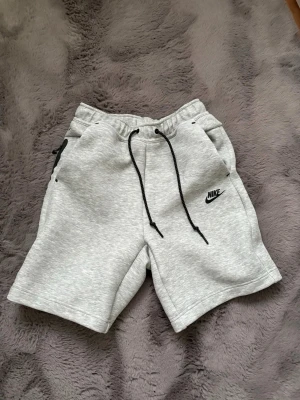 Gråa Nike tech shorts - Snygga ljusgrå shorts från Nike med svart snörning i midjan och dragkedjeficka bak. Mjukt material i bomullsmix. Andvänd 2 gånger och tvättad 1 gång annars inget annorlunda. Om du har några funderingar så kan du gärna medela mig