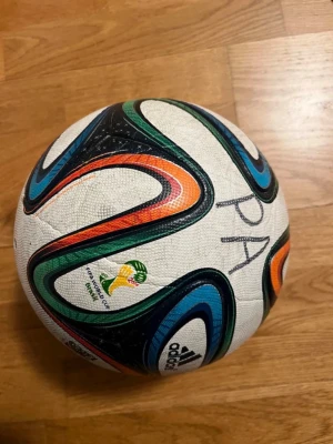 Adidas Brazuca fotboll VM 2014 - Säljer en Adidas Brazuca fotboll från VM 2014 i Brasilien. Bollen är vit med färgglada mönster i blått, grönt, orange och svart. Tillverkad i slitstarkt syntetmaterial och har officiell VM-logga tryckt på sidan.