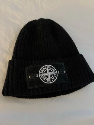 Svart ribbstickad mössa Stone Island - Svart ribbstickad mössa från Stone Island med uppvikt kant och den klassiska logotypen framtill. Märket sitter på en patch som är fastsatt med två knappar. Perfekt för kalla dagar och en stilren streetwear-look. Endast använd vid fåtal tillfällen.