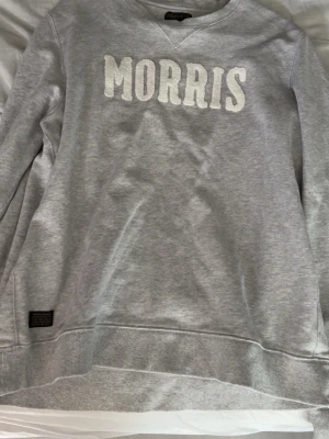 Grå sweatshirt från Morris - Snygg grå sweatshirt från Morris med vit broderad logga på bröstet. Tröjan har rund halsringning och ribbade muddar vid ärmslut och nederkant. Perfekt för en avslappnad och stilren look.
