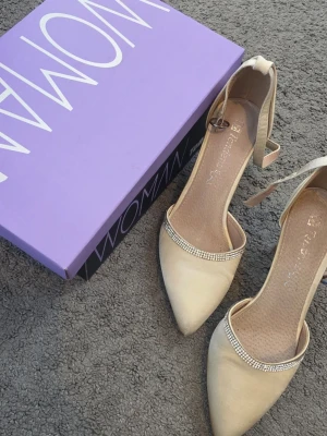 Beige klackar med strassdetalj - Snygga beige klackar med spetsig tå och glittrande strassdetalj över foten. Skorna har en smal ankelrem med spänne och är tillverkade i ett mjukt material som ger en elegant look. Storlek 37