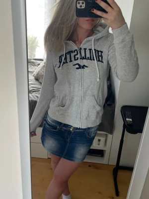 Grå Hollister hoodie med dragkedja - Snygg grå hoodie från Hollister med dragkedja framtill och stor broderad logga i mörkblått på bröstet.🥰