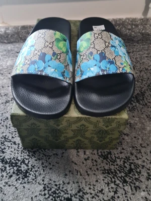Gucci sandaler med blått blommönster - Snygga sandaler från Gucci med svart sula i strukturmönstrat material och bred rem med klassiskt GG-mönster och blåa blommor. Perfekt för dig som vill sticka ut med exklusiv design och färgglada detaljer. Remmen har inslag av grönt och beige för en lyxig känsla.