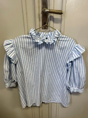 Blåvit randig blus med volanger - Söt blus med blå och vita vertikala ränder, volangdetaljer vid axlar och hals samt knäppning framtill. Blusen har puffiga långärmar med resår vid ärmslut och en hög krage med volang. Storlek 42 men funkar även 38