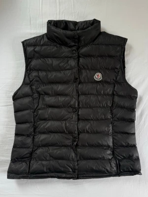 Moncler Vest - Snygg svart dunväst från Moncler med hög krage och klassisk logotyp på bröstet. Västen har horisontella sömmar och stängs med knappar framtill. Perfekt för lager-på-lager och håller dig varm med stil.