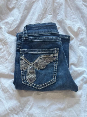 Blå jeans med broderad bakficka - Snygga blå jeans med cool broderad detalj på bakfickan. Jeansen har kontrastsömmar och klassisk femficksdesign. Perfekta för dig som vill ha ett par jeans med lite extra attityd. Byxorna är aldrig använda och är low waist🤗