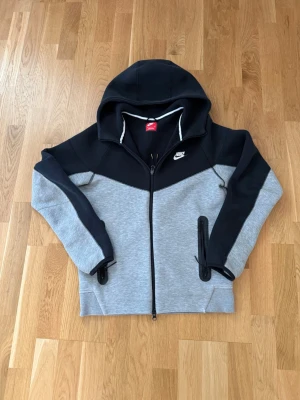 Nike hoodie i grått och svart - Snygg hoodie från Nike i grått och svart med dragkedja framtill och två fickor med svarta detaljer. Klassisk Nike-logga på bröstet och stor huva. Tillverkad i mjukt sweatshirtmaterial, perfekt för chill eller träning.