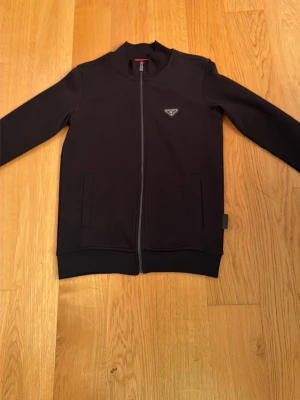 Svart zip-hoodie från Prada - Snygg svart hoodie från Prada med dragkedja framtill och klassisk logga på bröstet. Modellen har ribbade muddar och två fickor på sidorna. Tillverkad i ett mjukt och bekvämt material som passar perfekt till streetwear-stilen. 