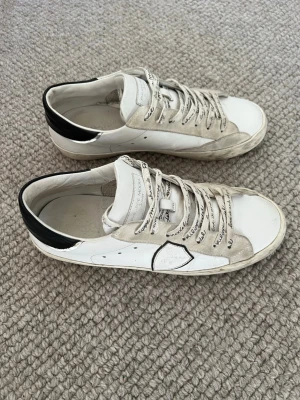 Philippe Model vita sneakers - Snygga vita sneakers från Philippe Model med svarta detaljer på hälen och textmönstrade skosnören. Skorna har en låg profil och är tillverkade i läder med inslag av mocka. Perfekta för dig som gillar en clean men ändå unik look. BOX INGÅR