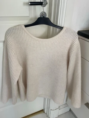 Stickad Gina tricot tröja med öppen rygg - Supersöt beige stickad tröja med rund hals och öppen rygg som knyts med en bred vit rosett. Tröjan har ribbstickat mönster och långa ärmar, perfekt för lager-på-lager och en trendig look.