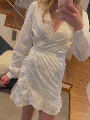 Vit omlottklänning med volang HELT NY! - Superfin vit omlottklänning från Bikbok med broderade blommor, långa ärmar och volangkant nertill. Klänningen har knyt i midjan och v-ringning, perfekt för sommarens alla tillfällen. Nypris 699
